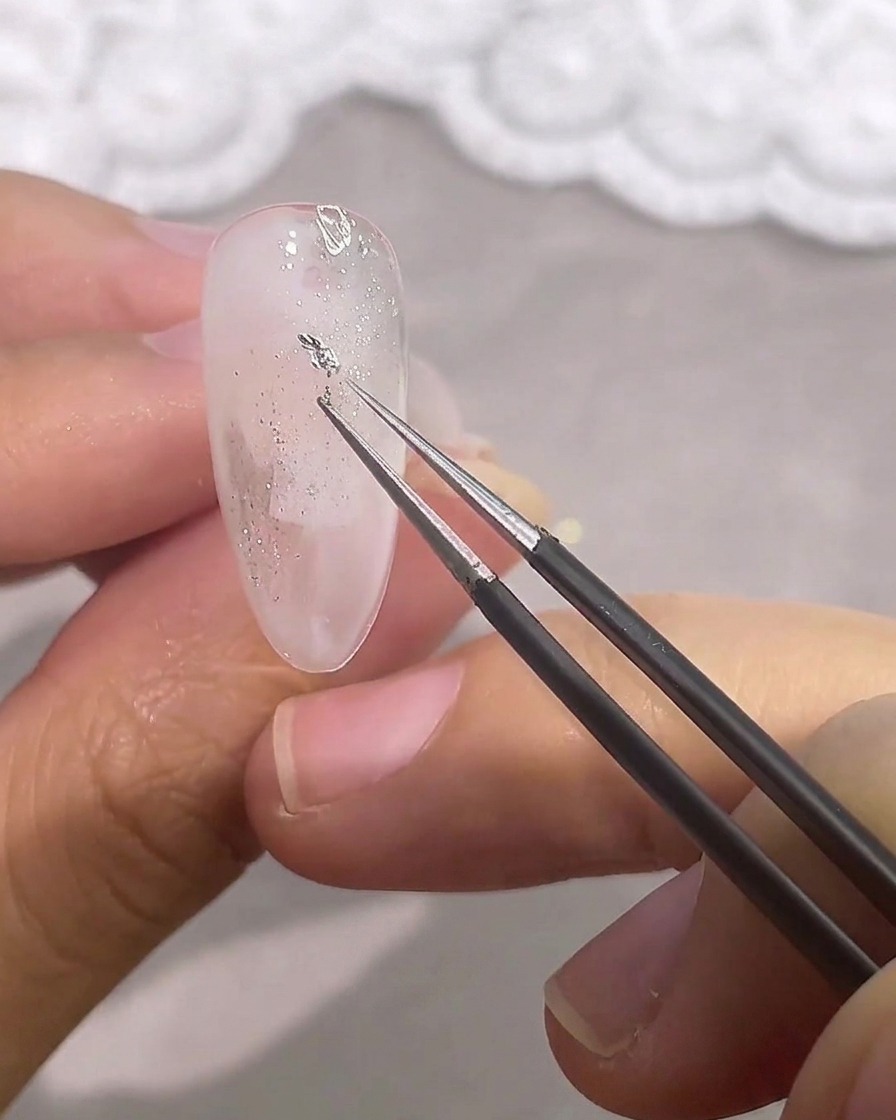 Delle pinzette posizionano un piccolo pezzo irregolare di foglia argento sullunghia.
