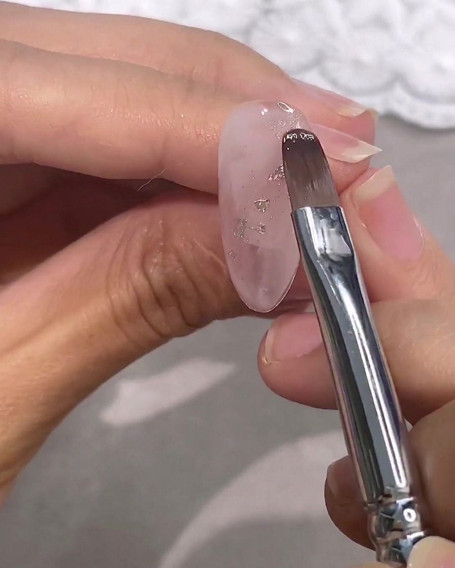 Un pennello stende un gel rosa semitrasparente sopra ununghia trasparente decorata con fiocchi argentati.