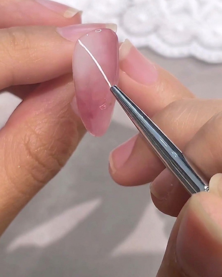 Un pennello fine per dettagli disegna una sottile linea diagonale bianca su ununghia in gel rosa.
