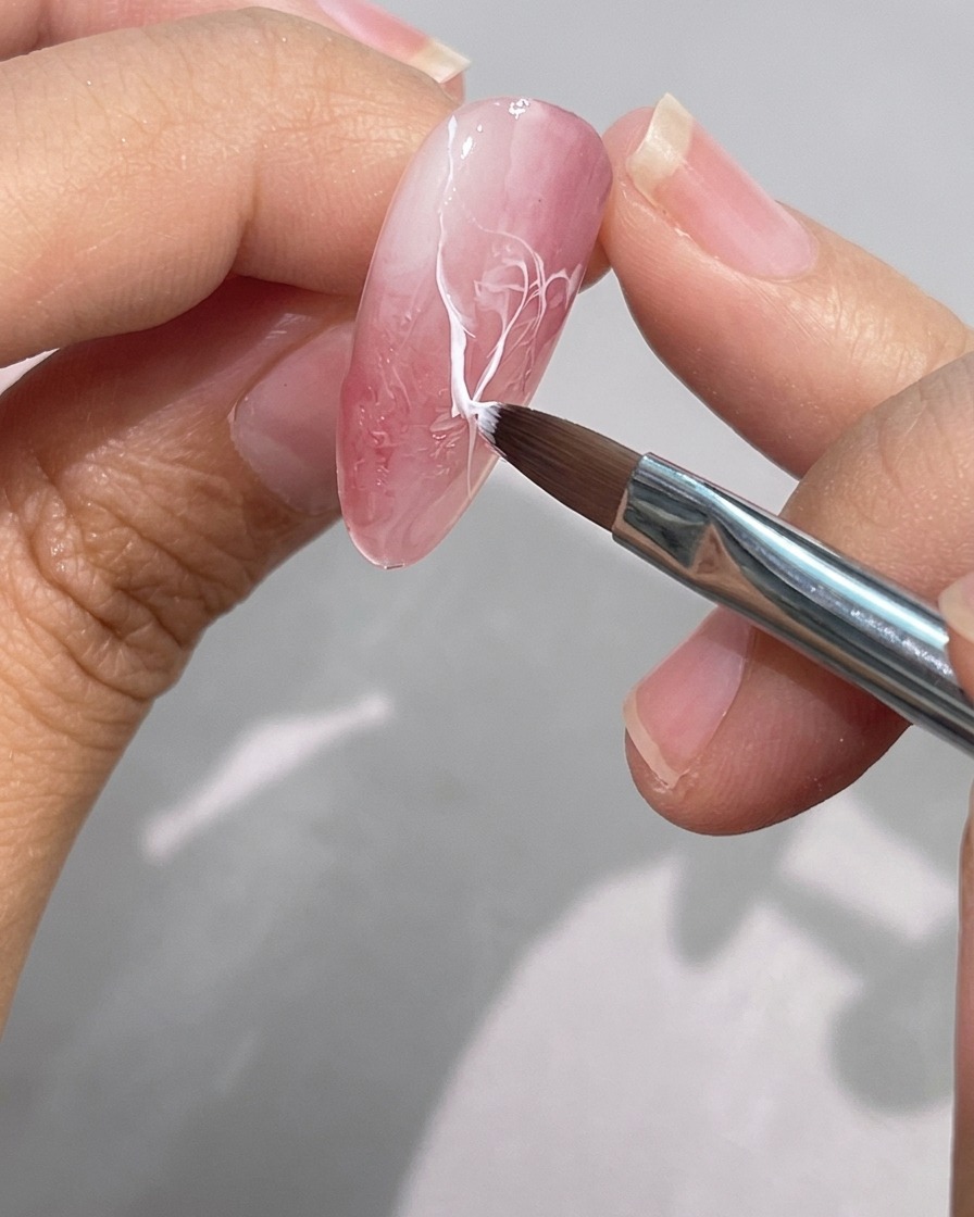 Un pennello fine per dettagli sfuma e ammorbidisce il bordo di una linea in gel bianco su ununghia effetto marmo rosa.
