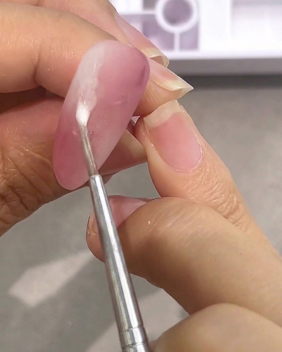 Un piccolo strumento applica una goccia di colore bianco acquoso su ununghia rosa opaca.