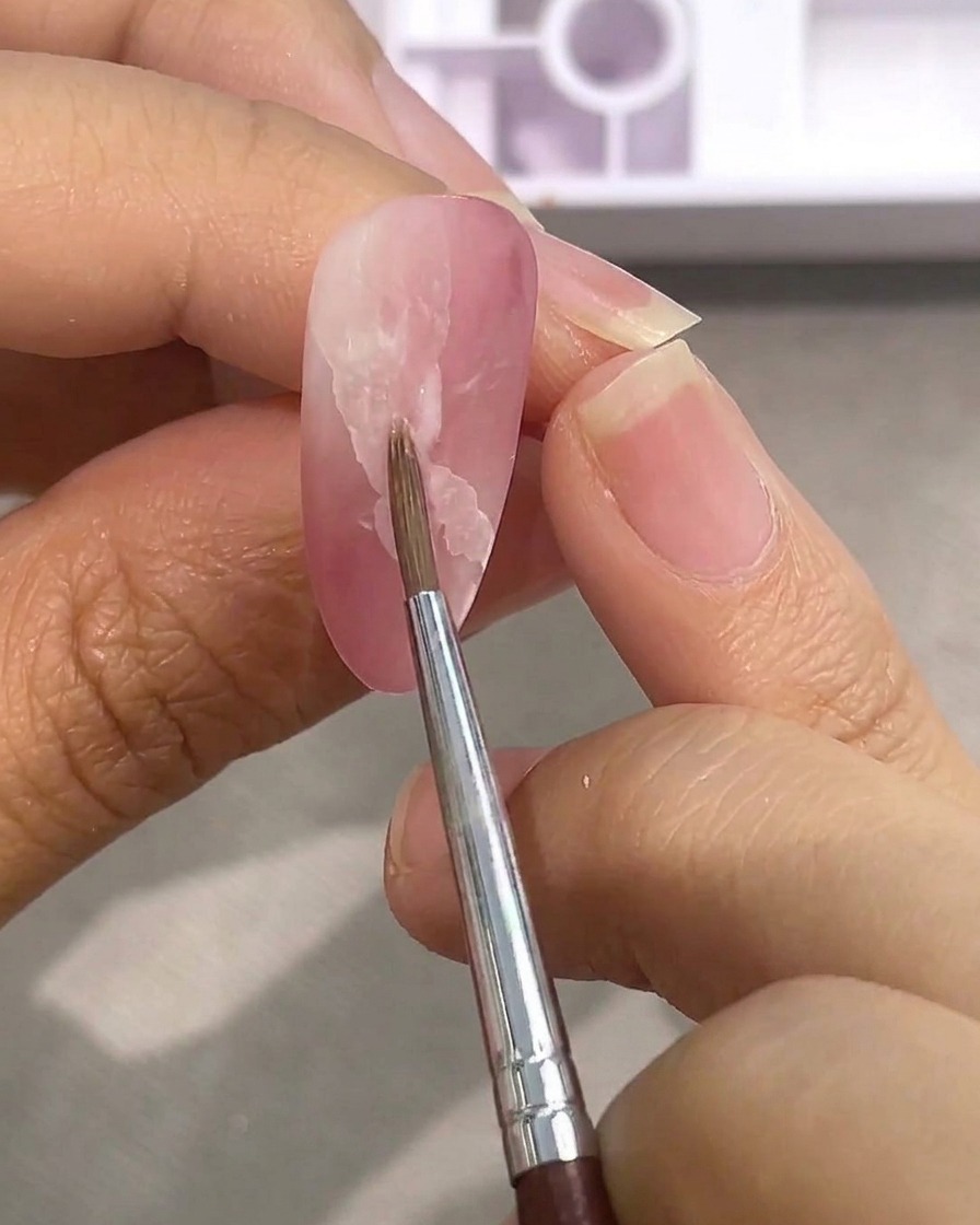 Un pennellino per dettagli sfuma i bordi di una macchia di acquerello bianco su ununghia rosa opaca utilizzando dellacqua.
