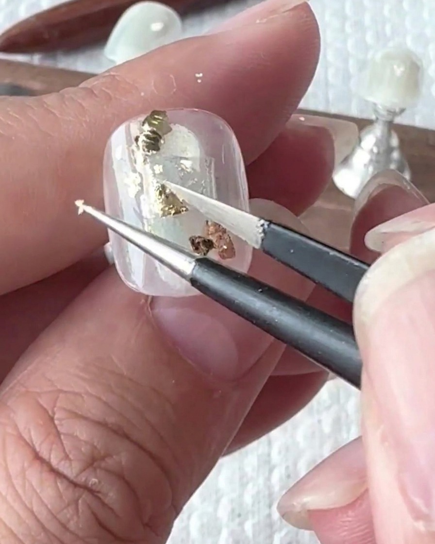 Pinzette che posizionano con cura pezzi irregolari di foglia oro sulla superficie dellunghia.