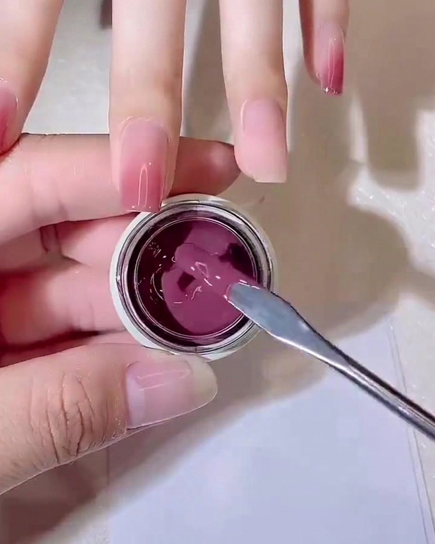 Una spatolina di metallo preleva una piccola noce di gel rosa-violaceo effetto jelly da un vasetto.