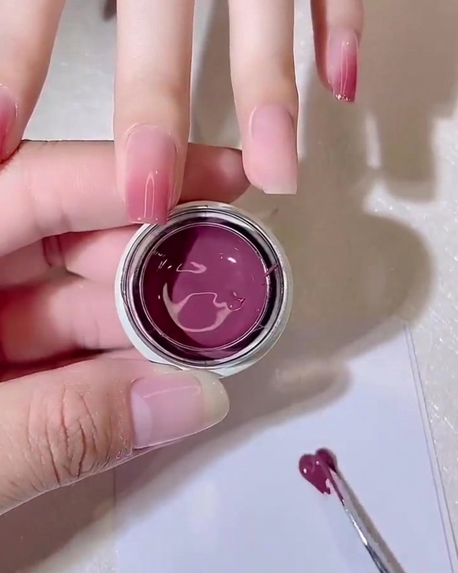 Uno strumento di metallo stende un sottile strato di gel rosa-violaceo su una palette di carta bianca.