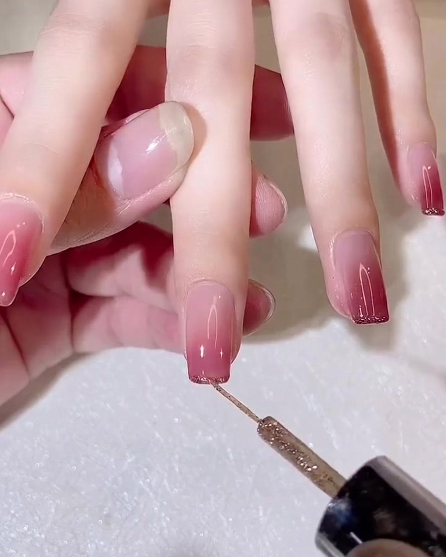 Un pennello a punta fine applica una sottile linea di gel glitter dorato sul margine libero di ununghia sfumata.
