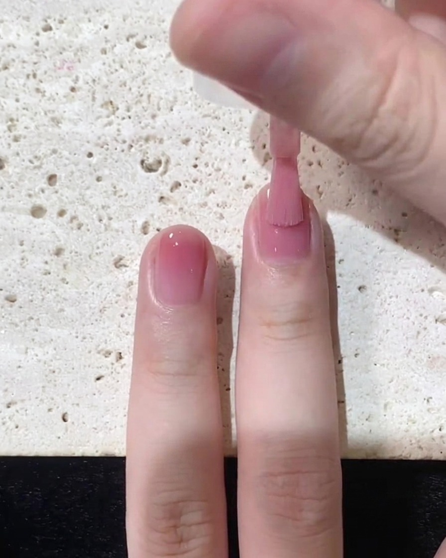 Un pennello che stende lo smalto jelly rosa fluidamente verso il bordo libero sulla metà superiore dellunghia