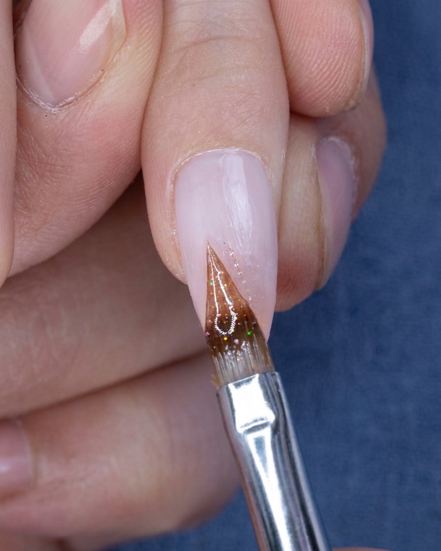 Primo piano di un pennello a punta sottile che applica piccoli glitter sulla punta di ununghia ombré.
