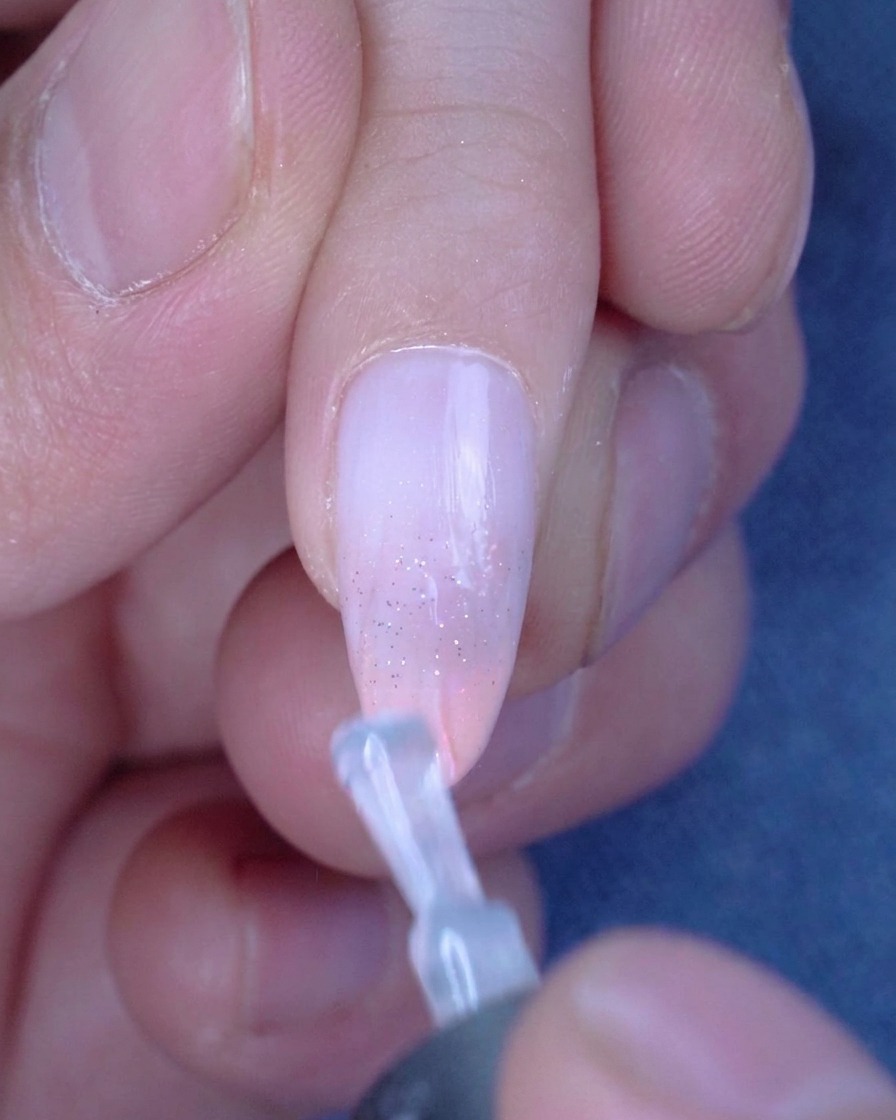 Un pennello applica uno strato lucido di top coat su ununghia ombré glitterata.