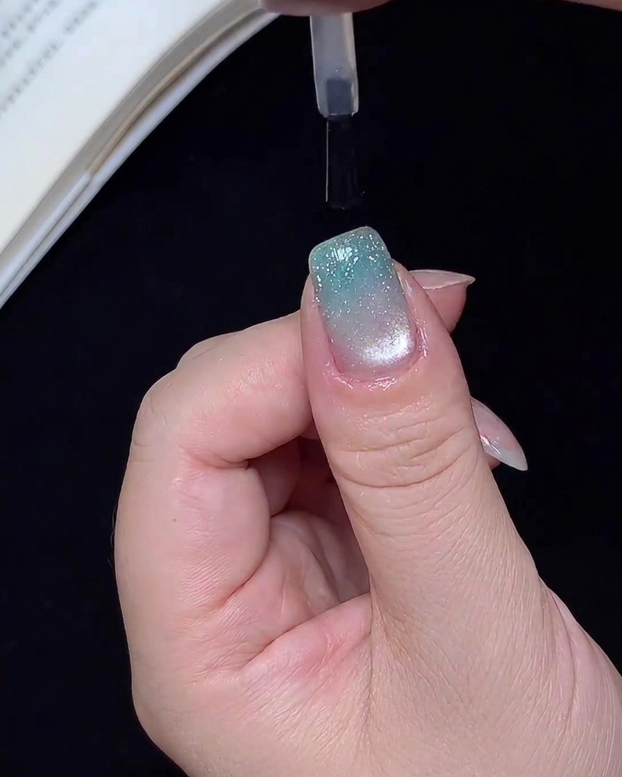 Un pennello che applica un top coat trasparente e lucido su ununghia con sfumatura glitter blu finita.
