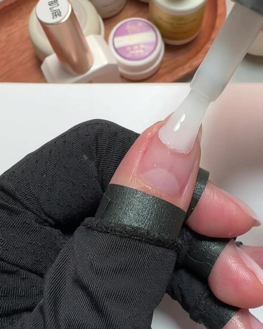 Primo piano di un pennello che applica smalto gel bianco latte su ununghia del pollice preparata.