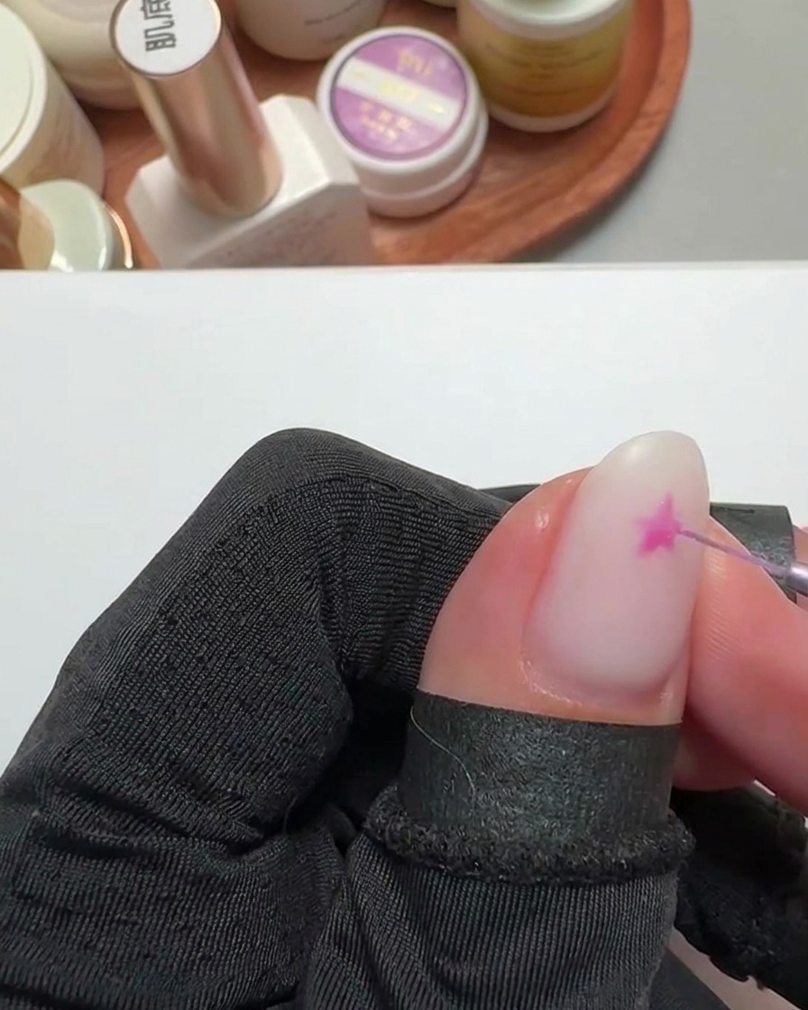 Primo piano di un pennello liner fine che disegna una stella rosa brillante su ununghia con base bianco latte.