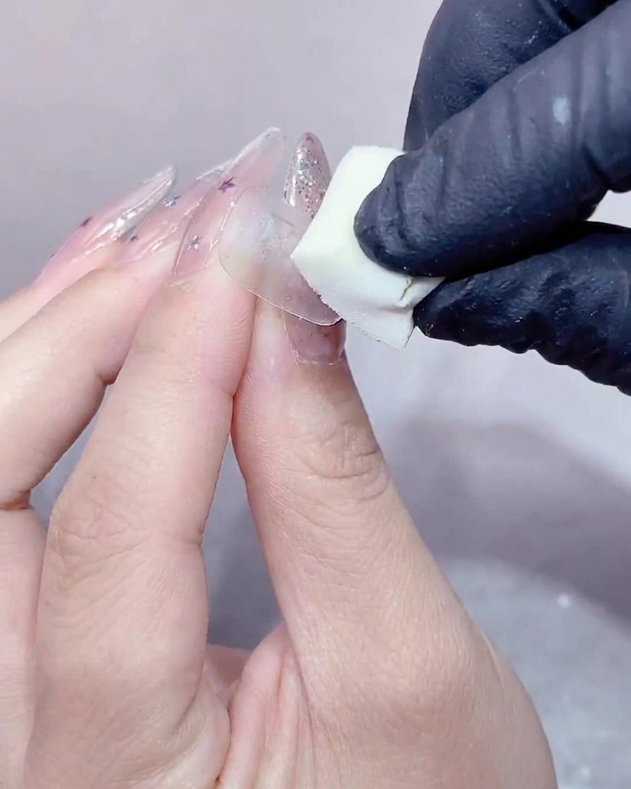Una mano con guanto che preme una piccola spugnetta da trucco bianca sulla superficie di ununghia ricoperta di gel trasparente.