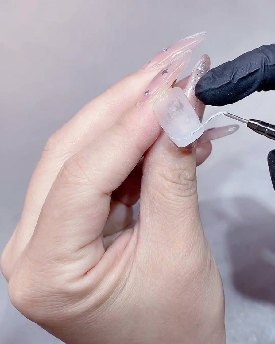 Un primo piano di unonicotecnica che utilizza un pennello piatto per riempire una punta french bianca su un allungamento in gel trasparente.