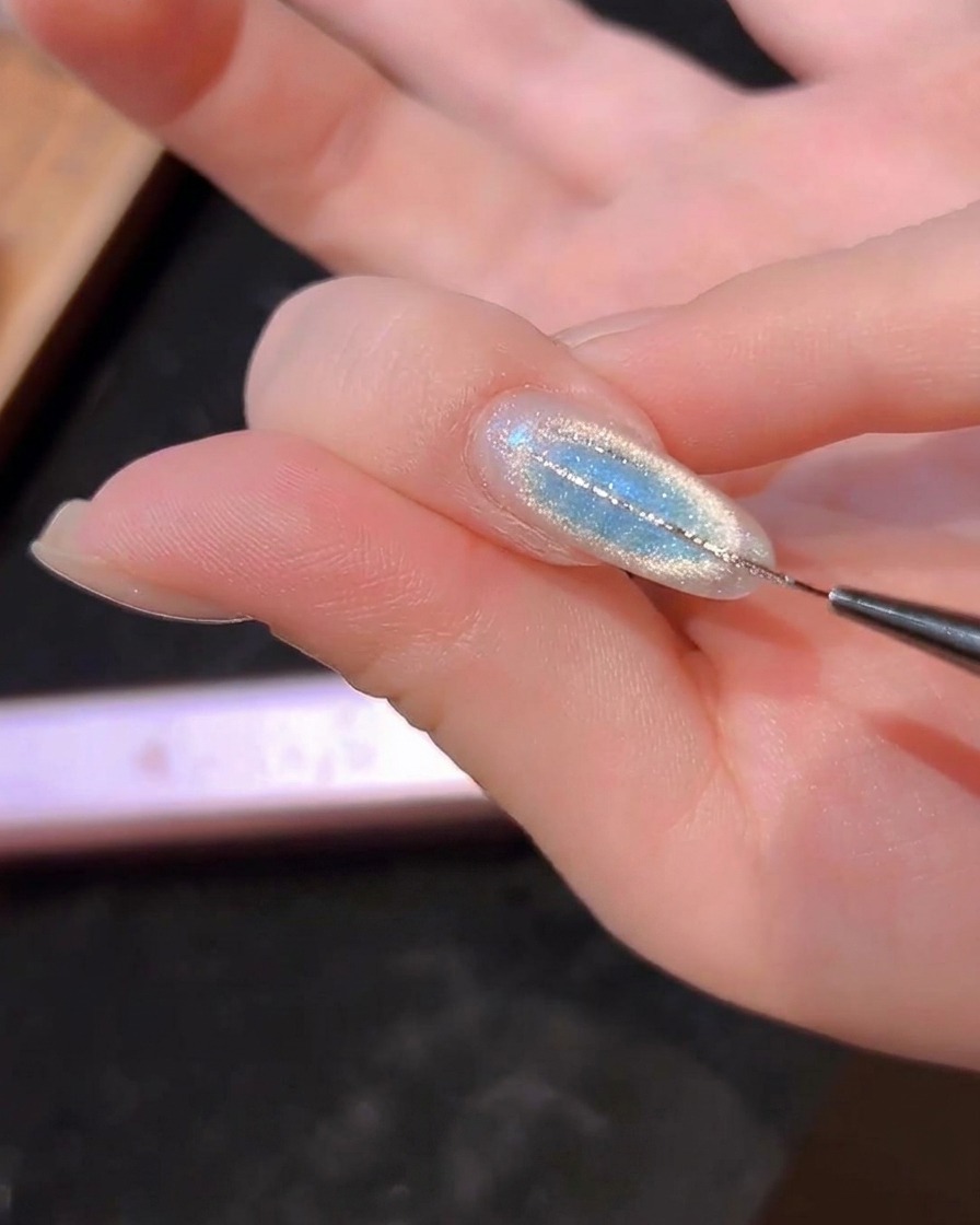 Un pennello liner sottile che disegna sottili linee argentate su ununghia cat-eye blu.