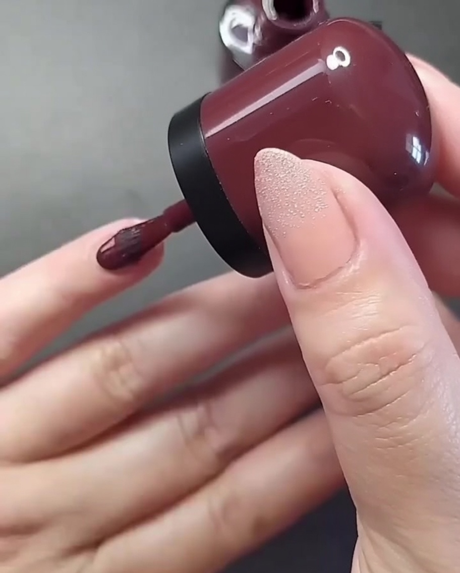 Primo piano di una mano con un pennello che applica smalto bordeaux intenso sullunghia del mignolo.