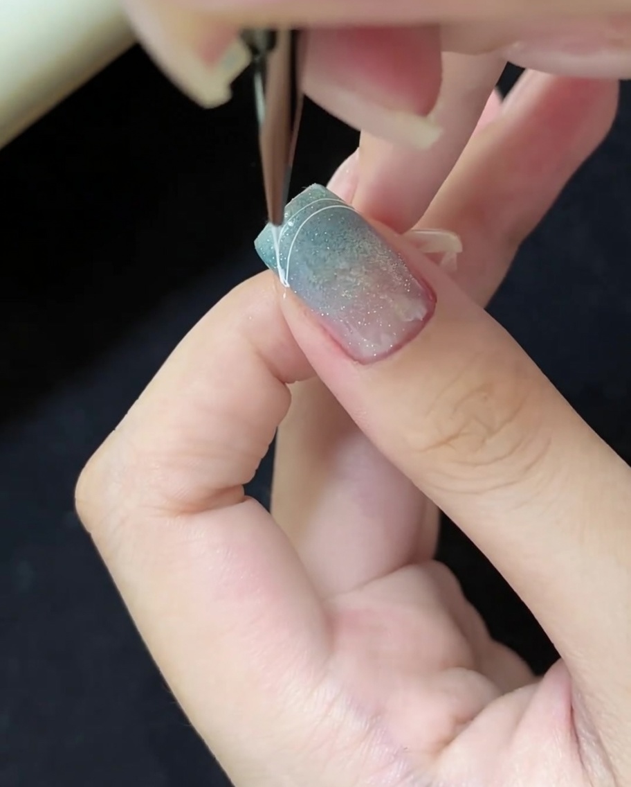 Primo piano di un pennello per unghie che disegna una sottile linea curva bianca sullangolo destro di una punta cat-eye blu.