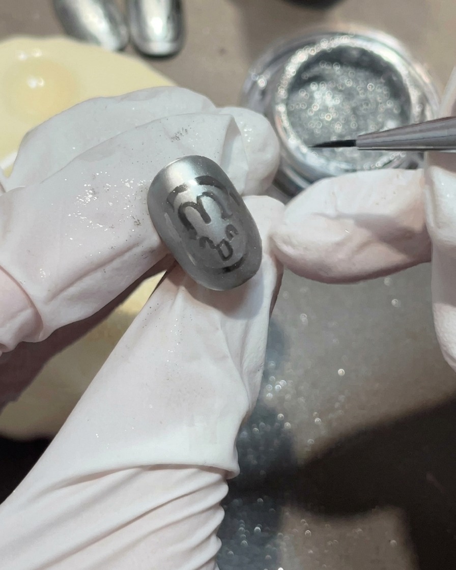 Dettaglio di un pennello liner che applica una piccola goccia di gel trasparente su ununghia grigio opaco.