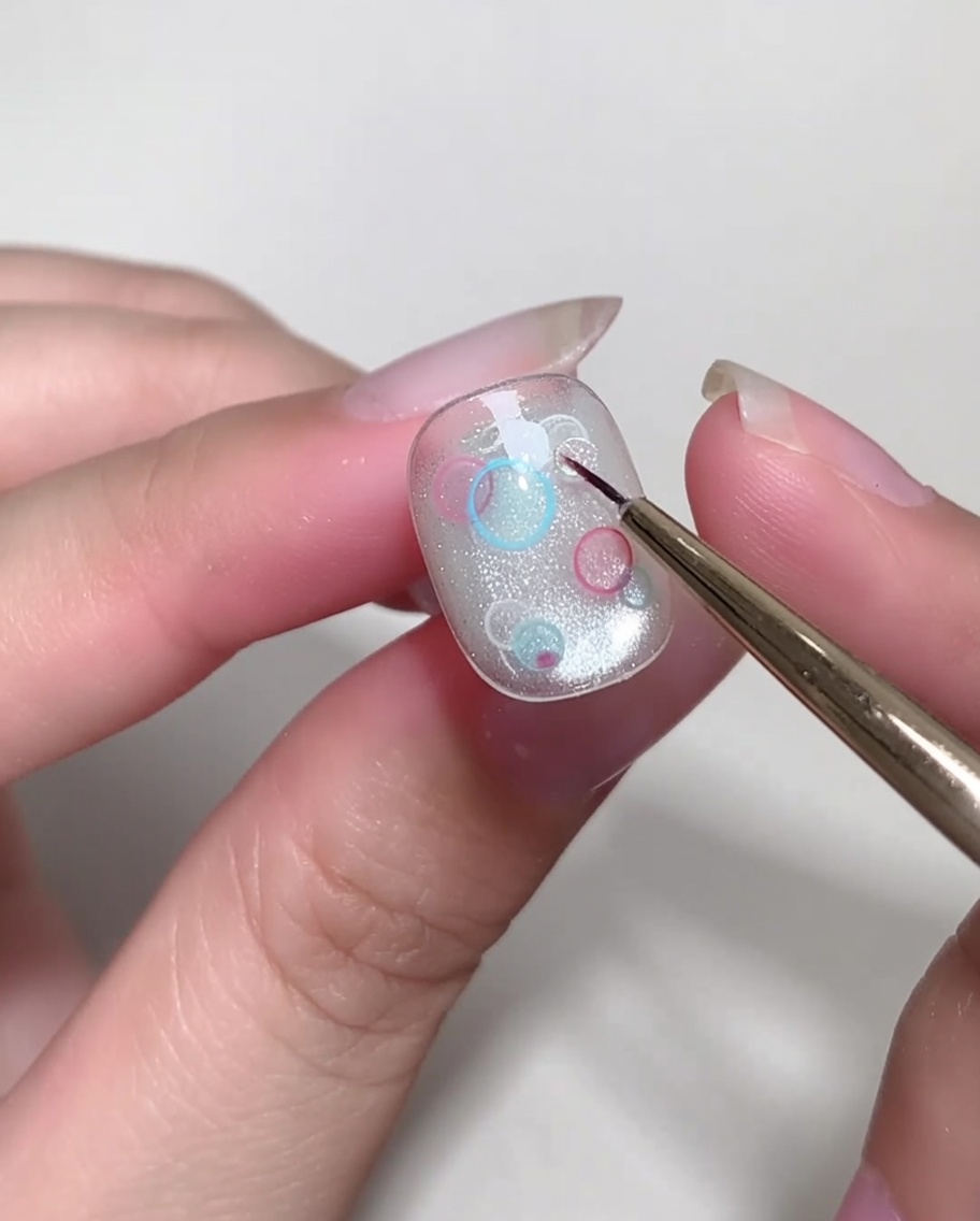 Un pennello a punta fine posiziona con cura piccole paillettes trasparenti e circolari sullunghia.