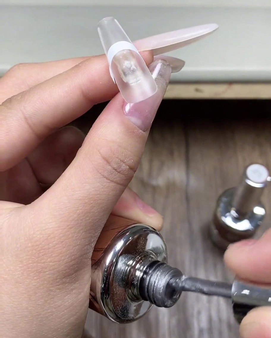 Un pennello che applica uno strato di gel cat eye grigio sulla punta di ununghia protetta da un adesivo.