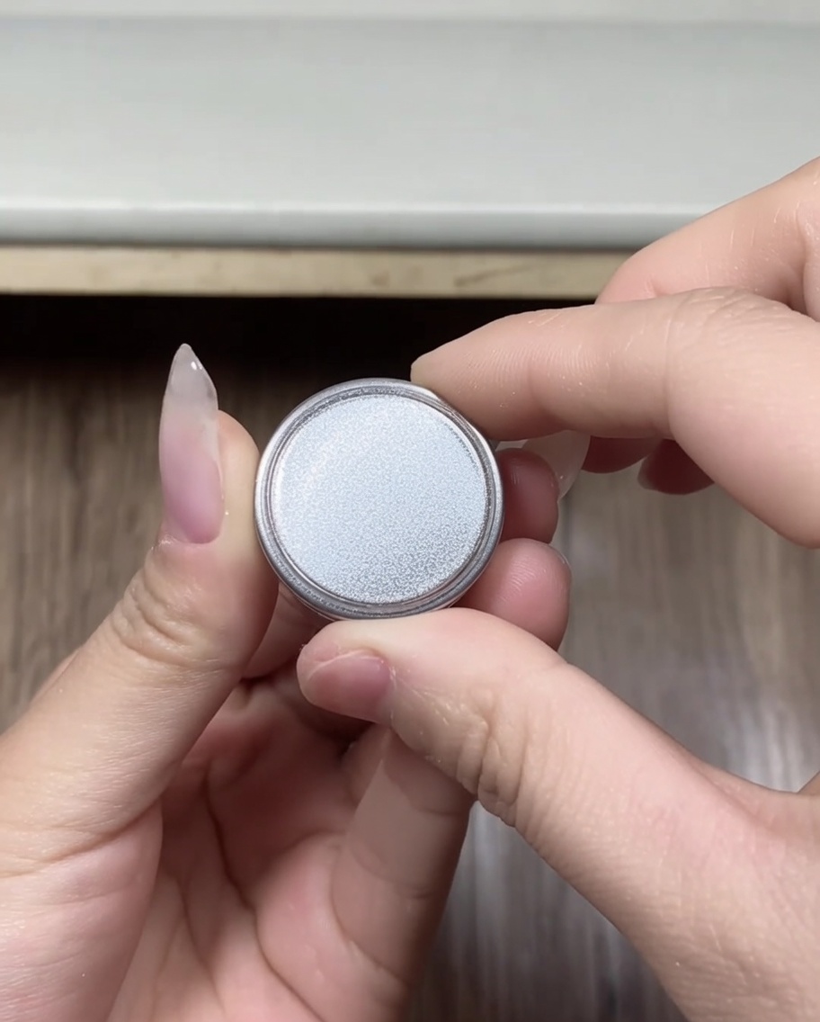 Primo piano di mani che tengono un piccolo contenitore rotondo di polvere cromata color argento, con il coperchio appena rimosso.