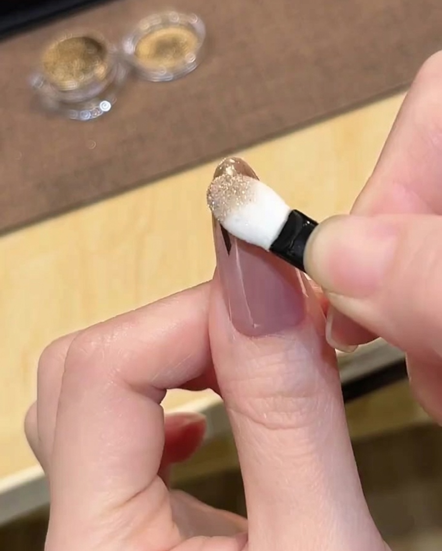 Un applicatore in spugna per ombretto che strofina la polvere cromata dorata sulla punta delimitata di ununghia.
