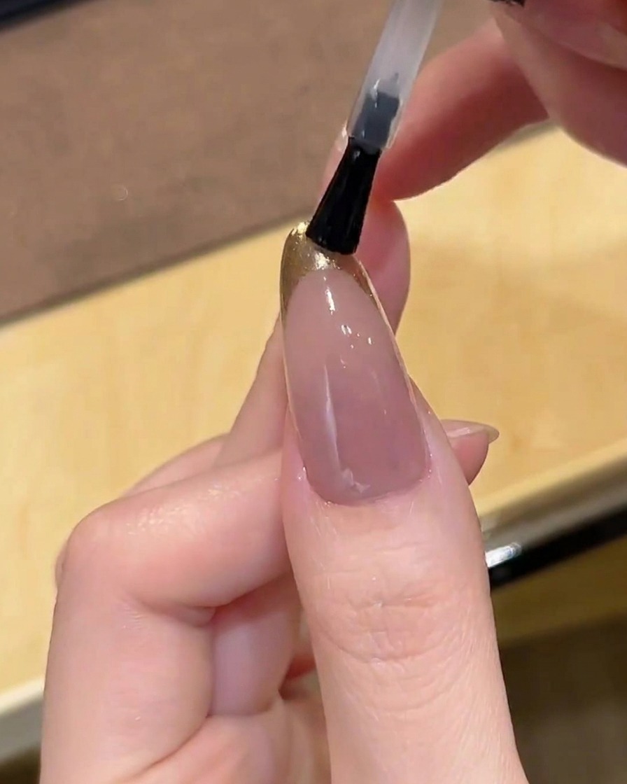 Un pennello che stende un top coat trasparente e lucido sopra ununghia con French cromato oro.