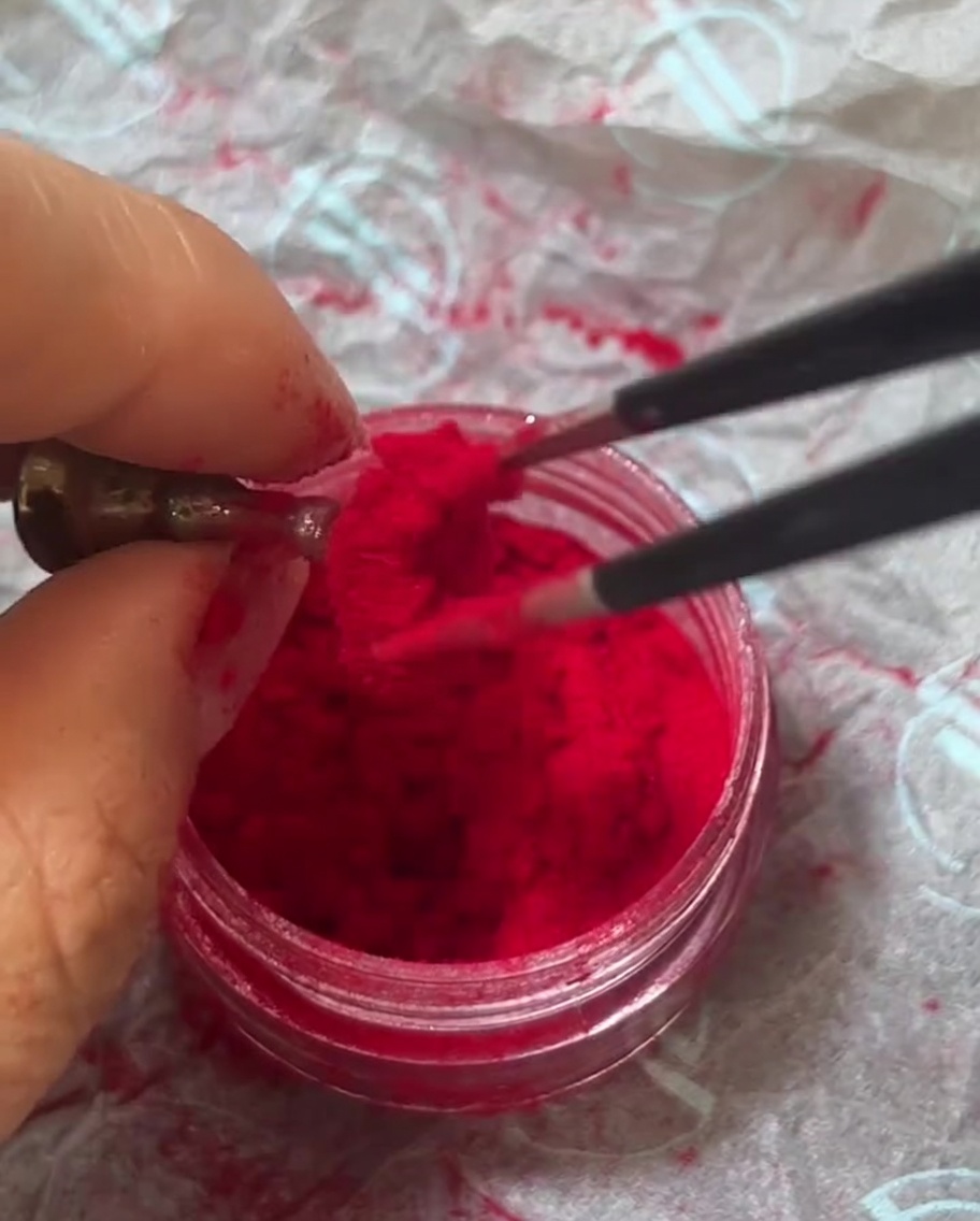 Pinzette tenute sopra un vasetto di polvere floccata rosso velluto che prelevano un ciuffo di fibre soffici.
