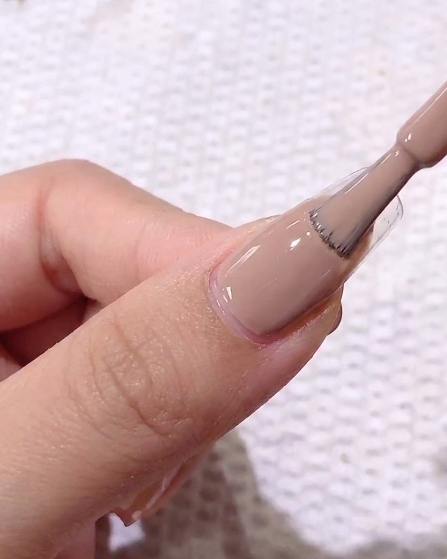 Primo piano di un pennello che applica uno smalto gel color marrone chiaro su ununghia naturale preparata con base trasparente.