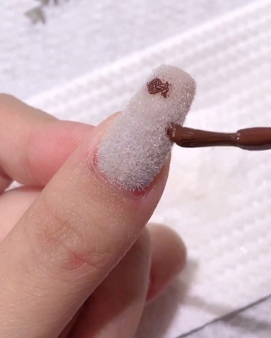 Un pennello da unghie che deposita un piccolo punto di gel color caffè scuro su una superficie opaca e vellutata bianca.