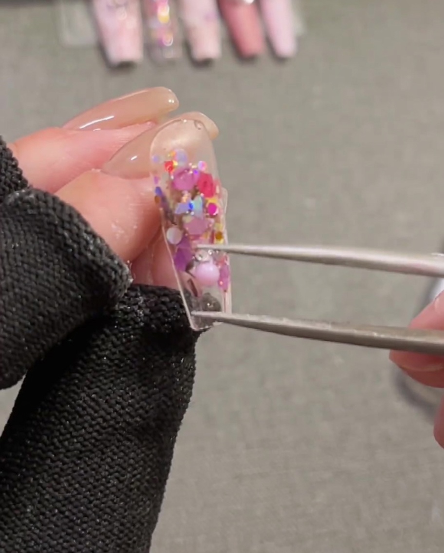 Pinzette posizionano un charm a forma di fiocco rosa 3D su ununghia con gel costruttore fresco.