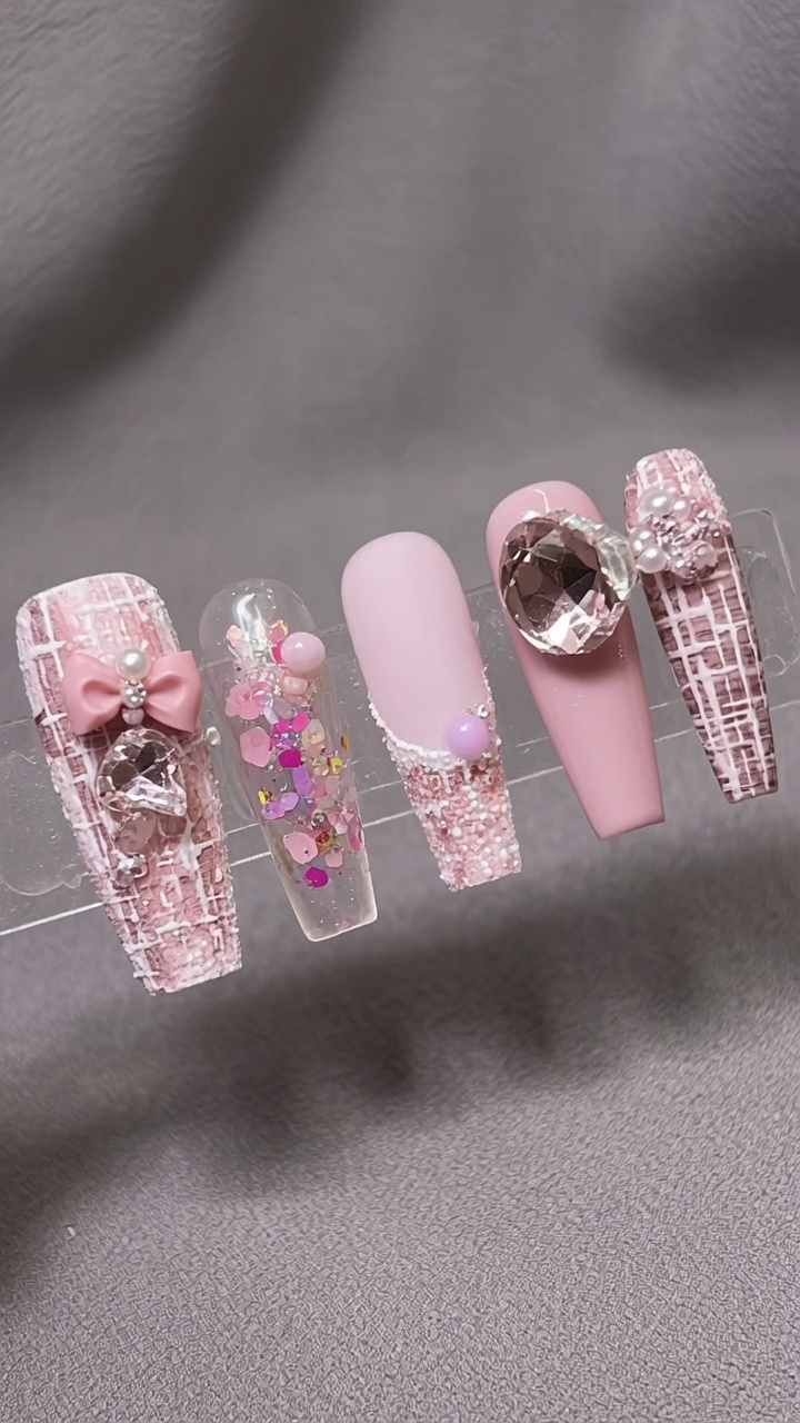Set di cinque unghie a stiletto con un tema invernale nei toni del rosa e bianco, caratterizzato da motivi tweed, paillettes iridescenti e decorazioni con fiocchi e gemme 3D.