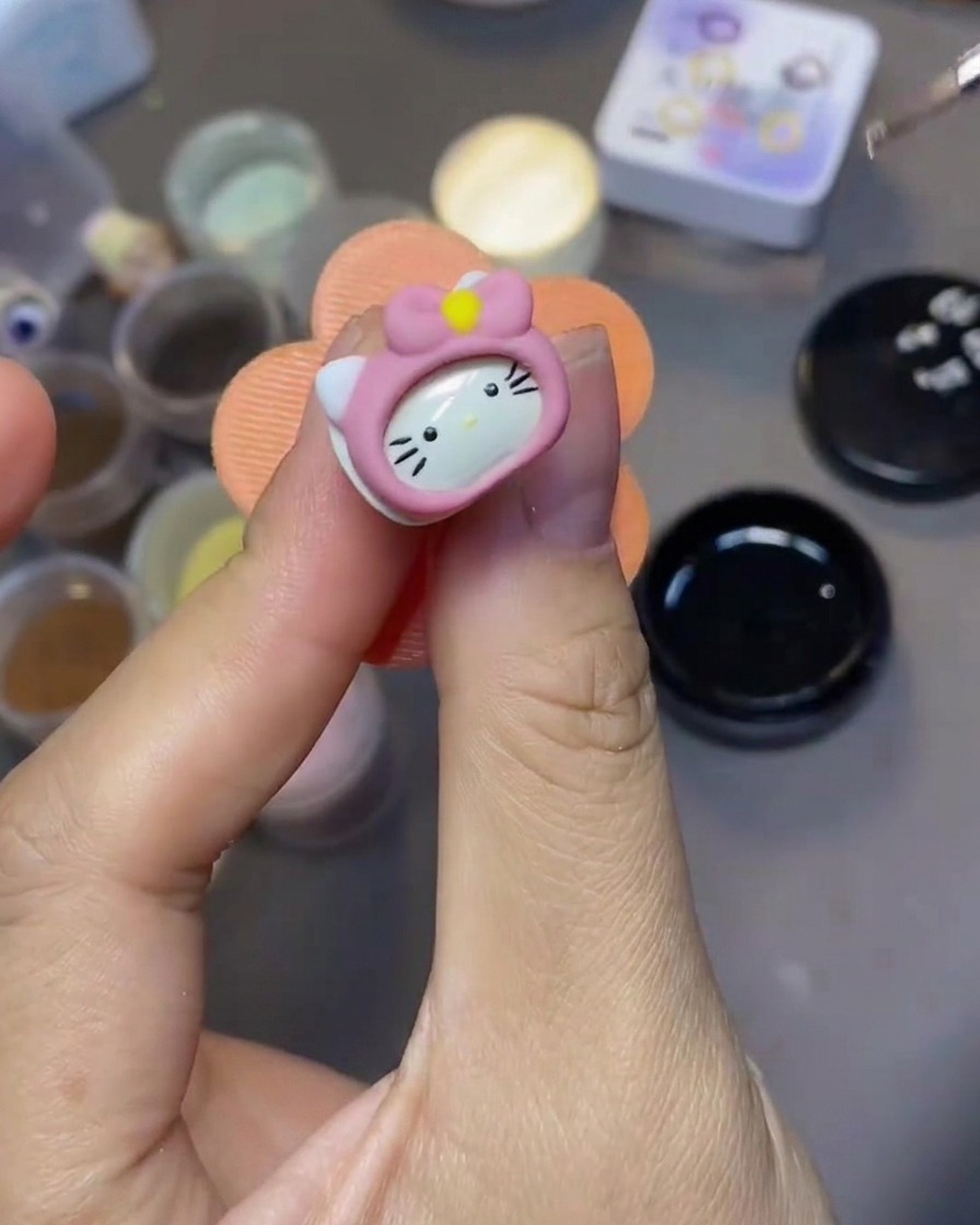 Un pennello fine da nail art applica un top coat trasparente su un charm 3D di Hello Kitty.