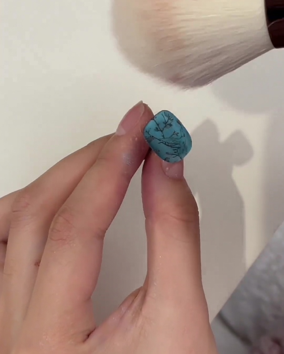 Un pennello da trucco che spolvera leccesso di polvere da ununghia turchese dopo la polimerizzazione.
