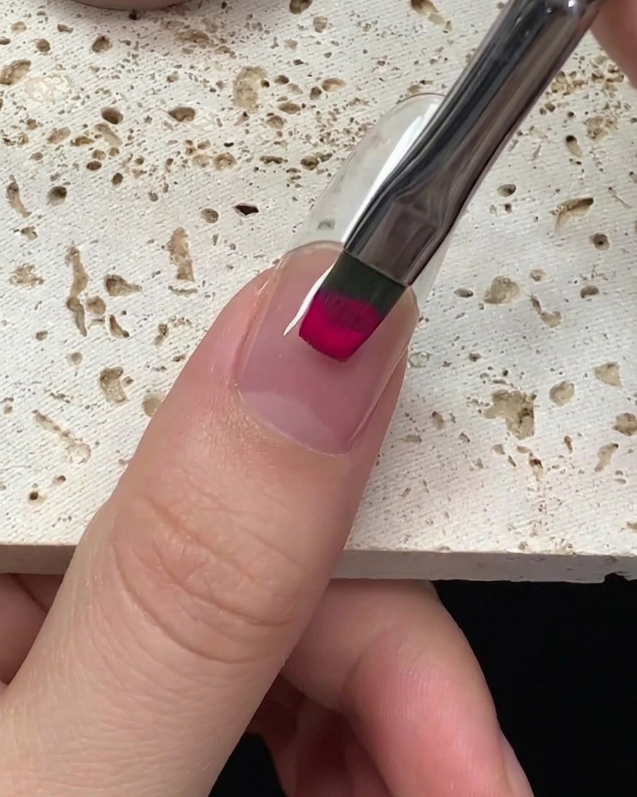 Primo piano di un pennello che applica smalto gel color magenta vibrante su ununghia, partendo dalle cuticole.