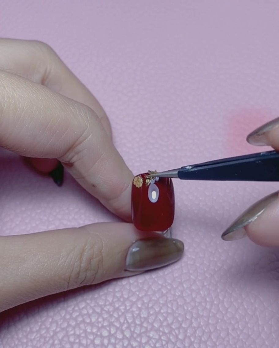 Un paio di pinzette che tengono un piccolo pezzo di foglia doro sopra ununghia laccata di rosso.