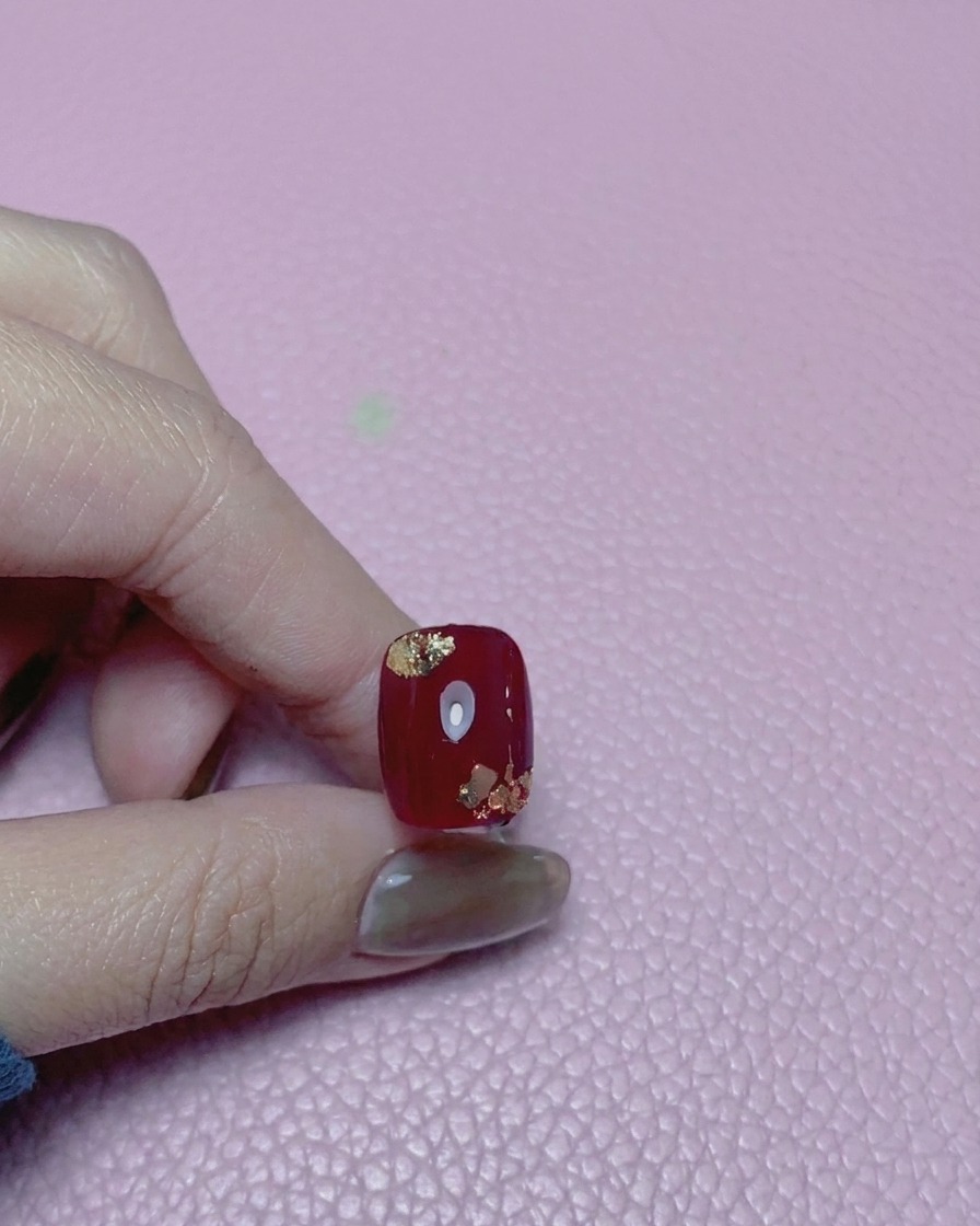 Primo piano di ununghia rossa con accenti in foglia doro, ricoperta da un top coat ultra lucido.