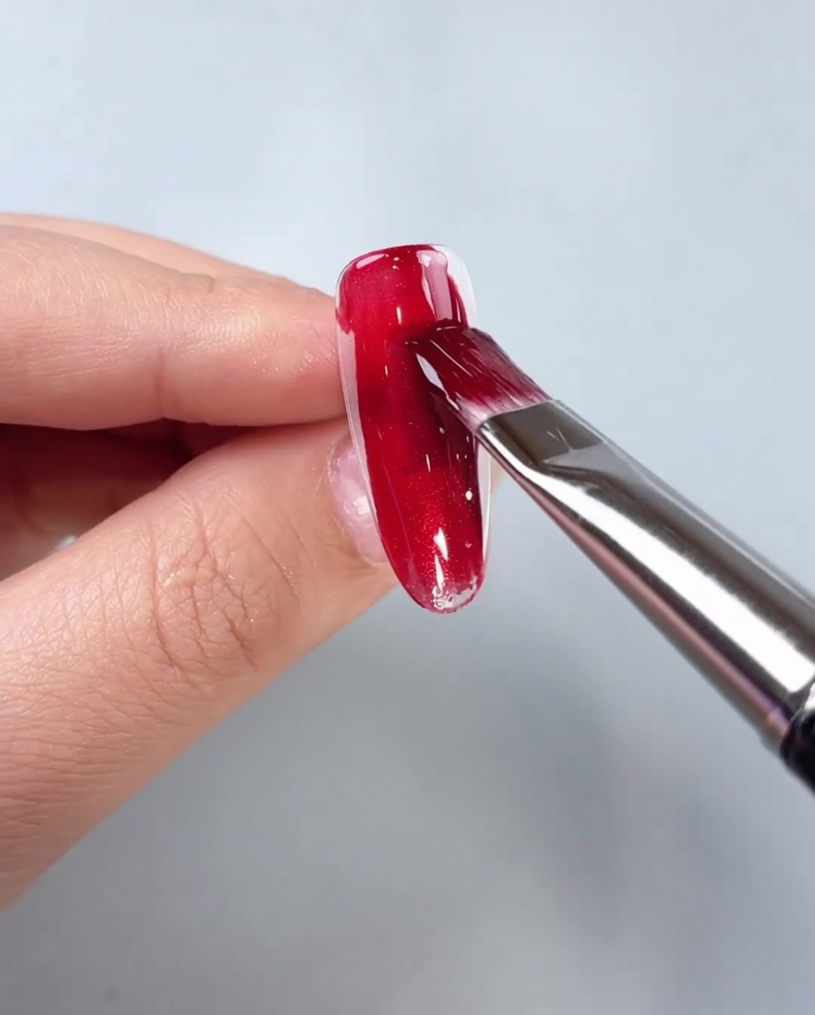 Un pennello applica il gel cat-eye rosso al centro di una tip per unghie trasparente.
