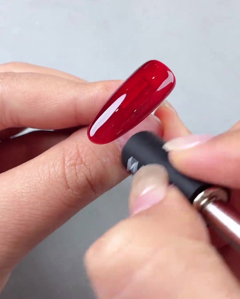 Un magnete per unghie tenuto ad angolo contro ununghia con gel cat-eye rosso per creare un effetto luminoso diagonale.