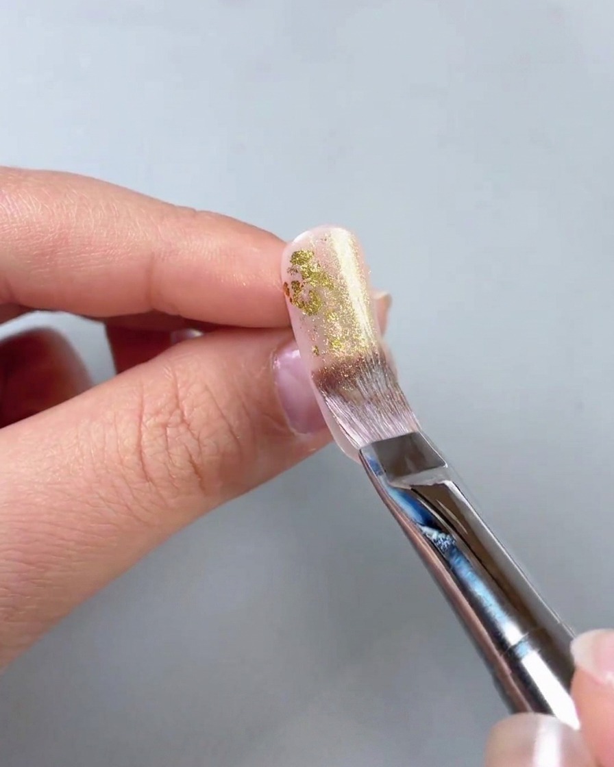 Primo piano di un pennello che applica un gel oro scintillante sopra frammenti di foglia oro.