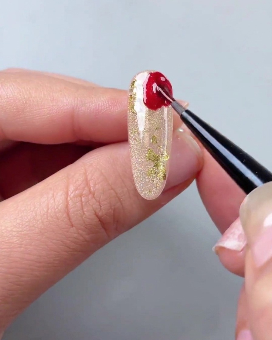 Un pennello di precisione applica gel cat-eye rosso in forme irregolari allangolo di ununghia oro.