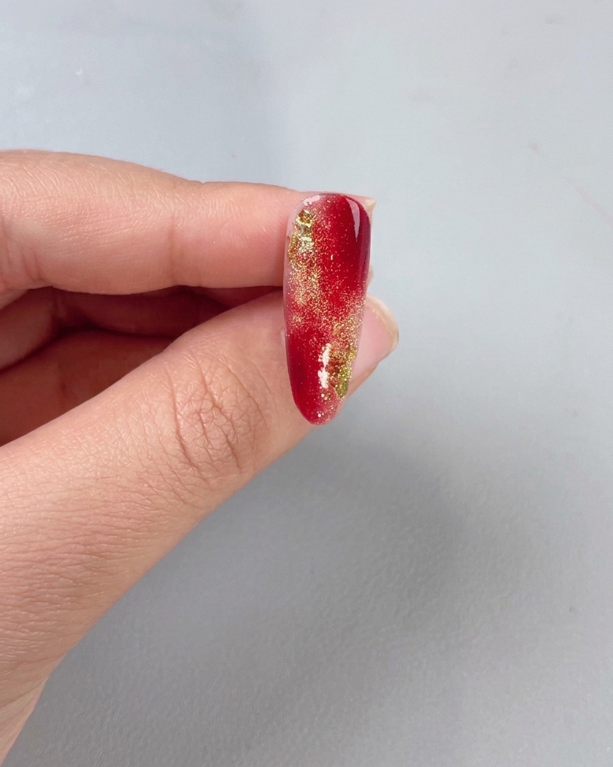 Un pennello applica un top coat trasparente sopra ununghia con gradiente rosso e oro.