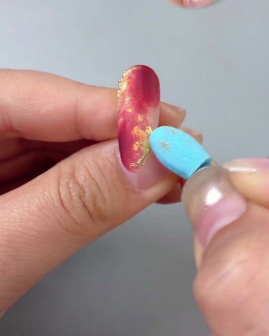 Primo piano di ununghia marmorizzata rosso e oro, mentre un applicatore a spugna rimuove la polvere oro in eccesso.