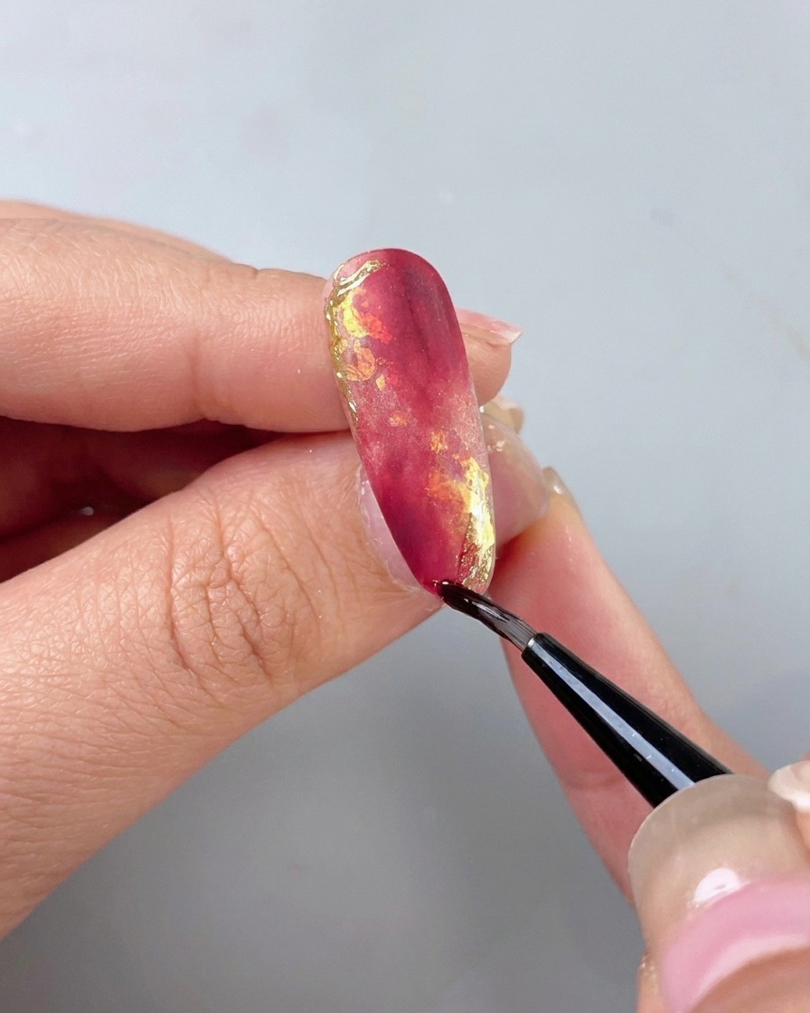 Un pennello sottile applica un top coat isolante trasparente sopra ununghia marmorizzata rosso e oro.