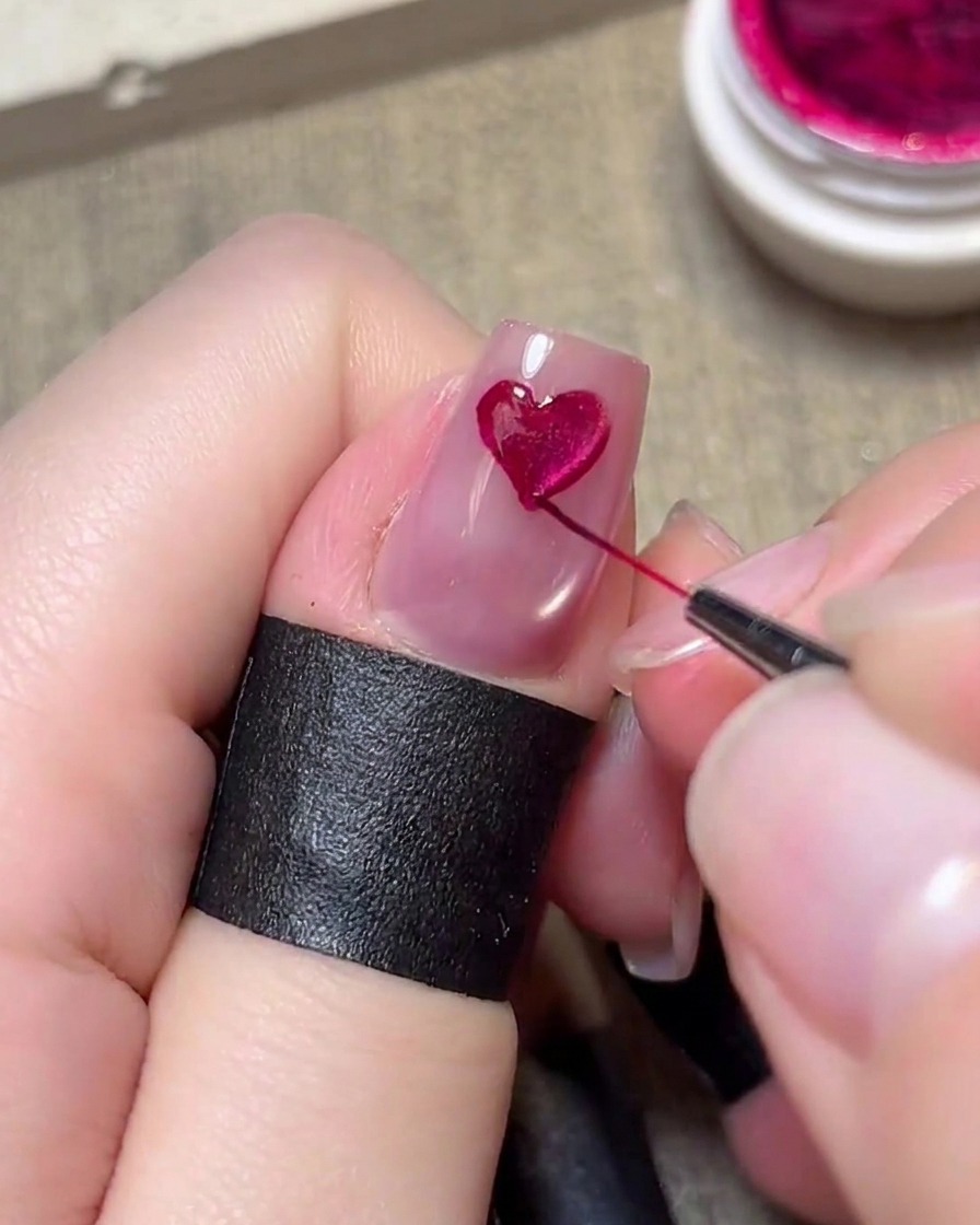 Un pennello a punta sottile che dipinge con cura un cuore magenta su ununghia rosa chiaro.