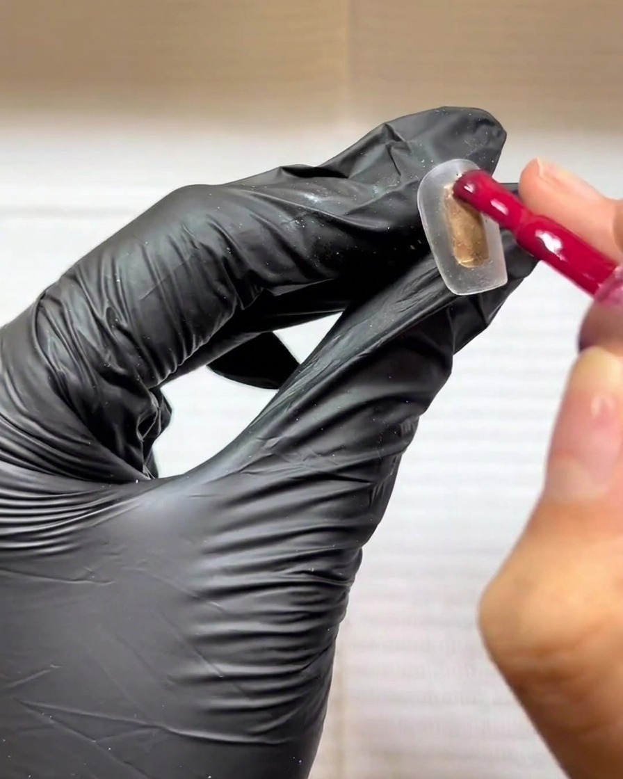 Una mano guantata utilizza un pennello per smalto gel per applicare un colore rosa-rosso su una tip trasparente.