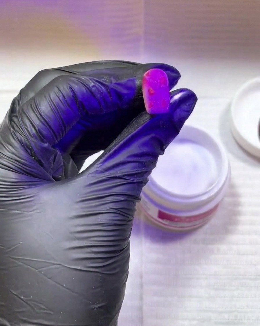 Una mano con guanto nero tiene ununghia rosa brillante con macchie texturizzate sotto una lampada UV/LED viola.