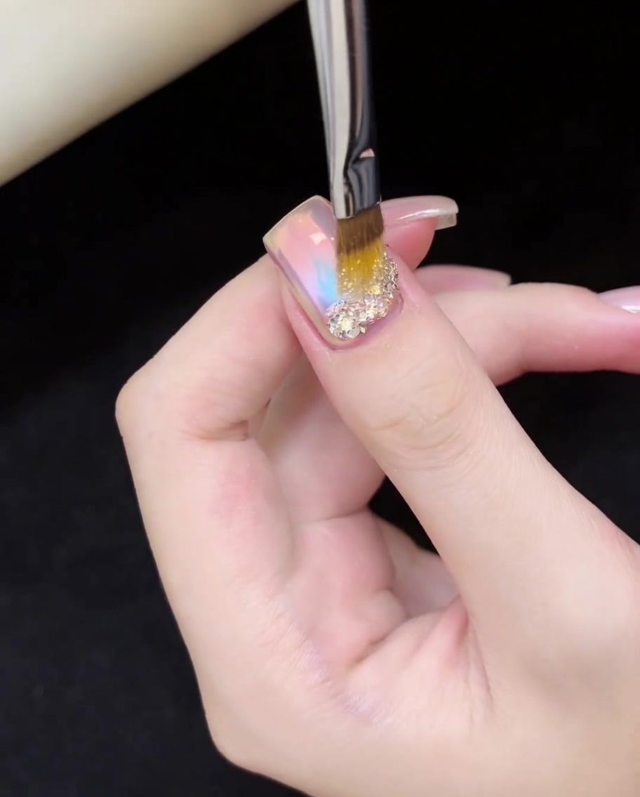 Un pennello per unghie che posiziona con precisione scaglie di glitter iridescenti oro e argento sulla base dellunghia.