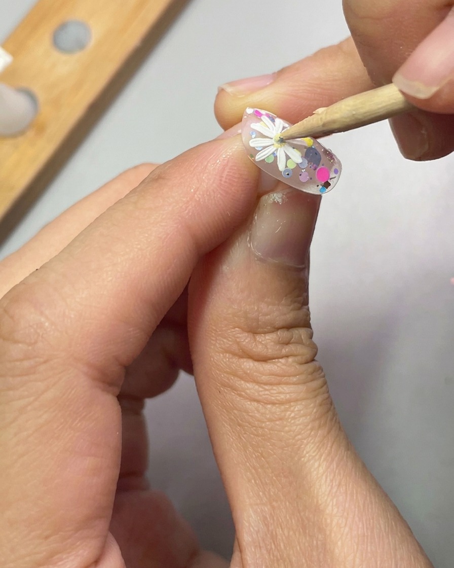 Uno strumento a sfera posiziona una piccola goccia di gel giallo al centro di una margherita bianca 3D su ununghia decorata.