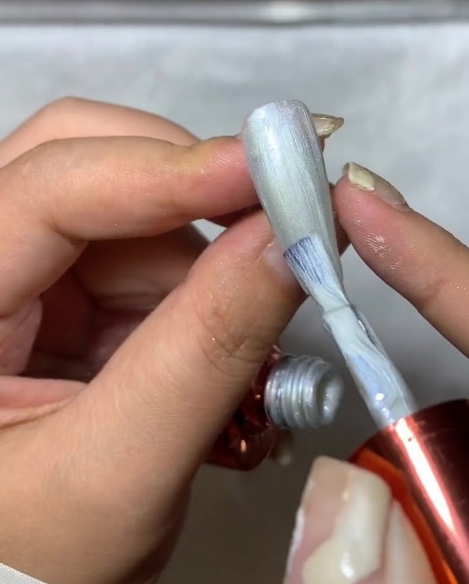 Un primo piano di un pennello da unghie che applica spider gel argento su ununghia, creando linee sottili e texturizzate simili a una ragnatela.